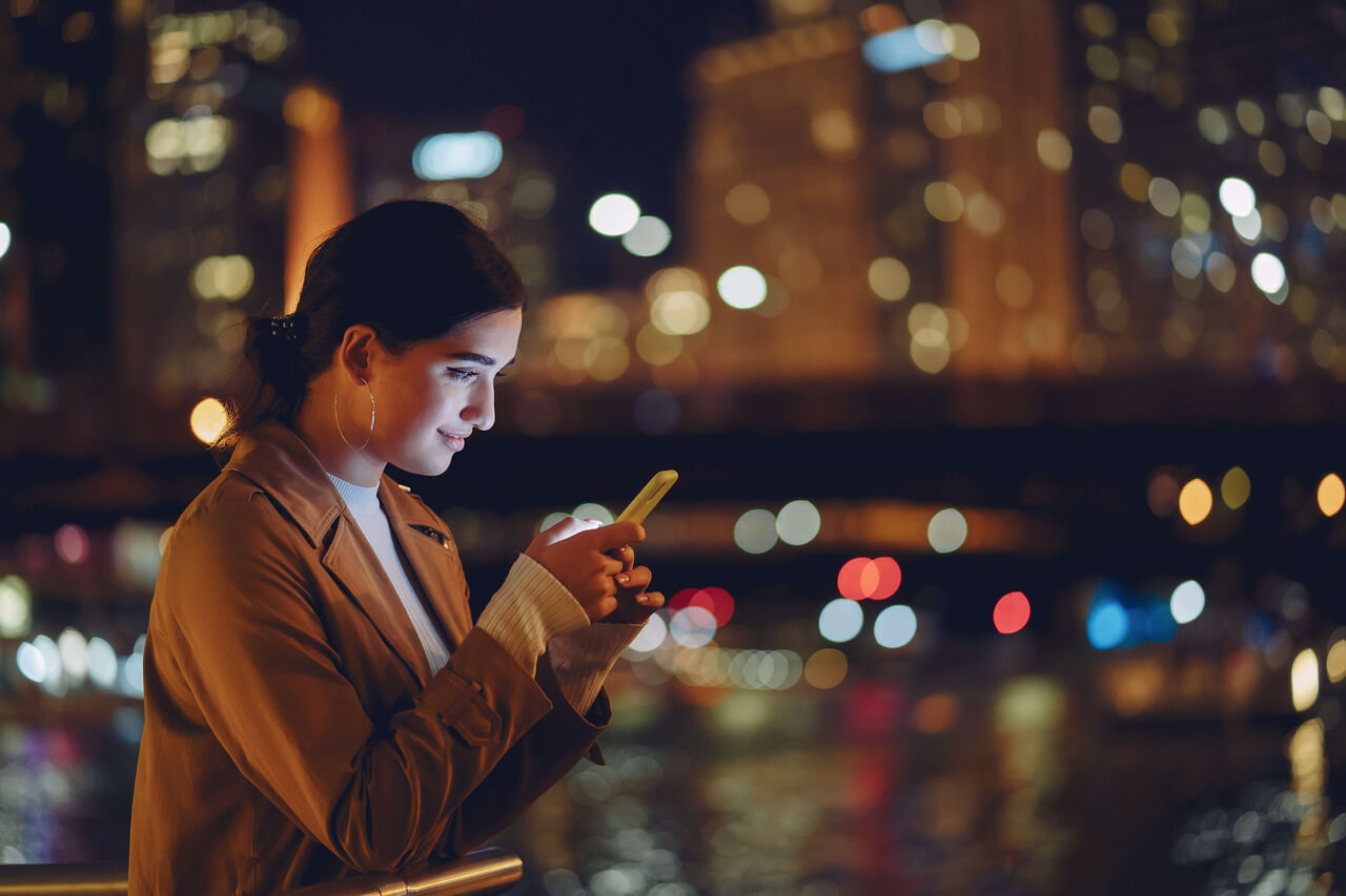 A imagem mostra uma jovem mulher usando um celular enquanto está ao ar livre à noite, em frente a um cenário urbano iluminado. Ela está com uma expressão concentrada, olhando para a tela do telefone, enquanto as luzes da cidade ao fundo criam um ambiente noturno vibrante e colorido. A iluminação das ruas e prédios proporciona uma atmosfera suave, com contraste entre as luzes quentes e o fundo escuro. 