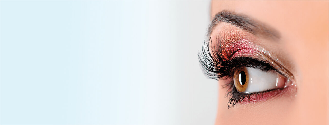 A imagem mostra o close-up de um olho feminino, com maquiagem vibrante nas cores dourado, vermelho e rosa, com brilho. Os cílios são longos e volumosos, destacando-se bem. A íris é marrom, e o olhar é direto, com foco nos detalhes da maquiagem e dos cílios. O fundo é suave e desfocado, destacando a área ao redor dos olhos.