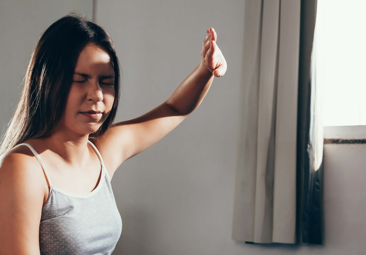 Mulher com os olhos fechados, levantando o braço direito à frente, como se estivesse pedindo para parar ou sinalizando algo. A luz natural entra pela janela ao fundo, iluminando suavemente o ambiente. A expressão dela é de concentração ou reflexão.