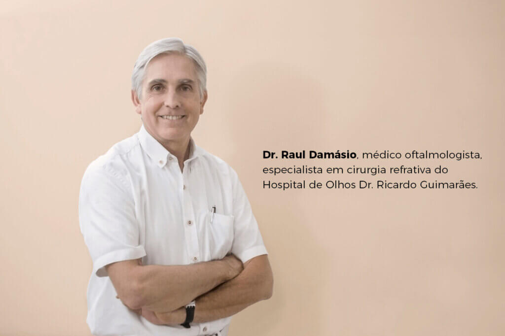 A imagem apresenta o Dr. Raul Damásio, médico oftalmologista, especialista em cirurgia refrativa, vestindo uma camisa branca e sorrindo enquanto está de braços cruzados. Ele está posicionado diante de um fundo de cor neutra, suave e clara. Ao lado dele, há uma legenda informando sua profissão e especialidade: "Dr. Raul Damásio, médico oftalmologista, especialista em cirurgia refrativa do Hospital de Olhos Dr. Ricardo Guimarães."