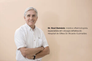A imagem apresenta o Dr. Raul Damásio, médico oftalmologista, especialista em cirurgia refrativa, vestindo uma camisa branca e sorrindo enquanto está de braços cruzados. Ele está posicionado diante de um fundo de cor neutra, suave e clara. Ao lado dele, há uma legenda informando sua profissão e especialidade: "Dr. Raul Damásio, médico oftalmologista, especialista em cirurgia refrativa do Hospital de Olhos Dr. Ricardo Guimarães."