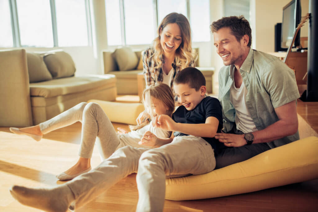 Imagem de uma família sorridente e feliz dentro de casa. Os pais, um homem e uma mulher, estão se divertindo com seus dois filhos, uma menina e um menino, enquanto brincam no chão. Todos estão sorrindo, e o ambiente parece acolhedor, com sofás e muita luz natural entrando pelas janelas, criando um clima alegre e familiar.