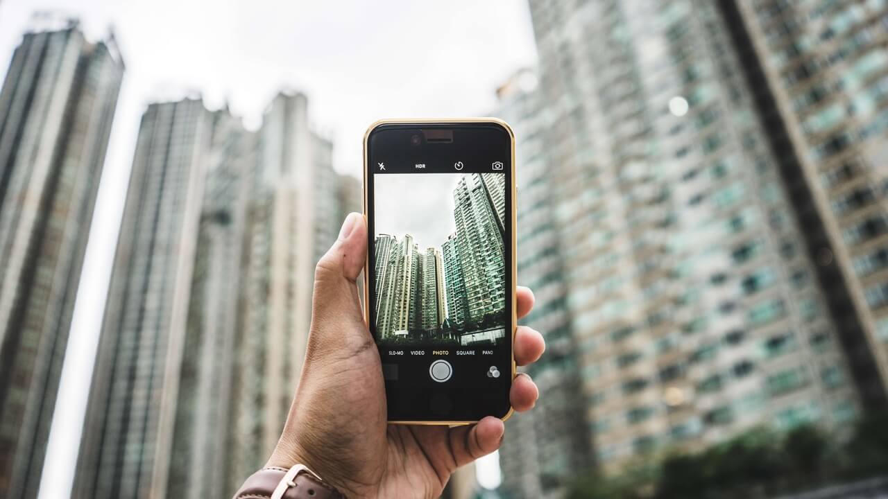 A imagem mostra uma pessoa segurando um celular em sua mão, com a tela do dispositivo exibindo uma foto tirada da vista de prédios altos. A foto no celular reflete a perspectiva de um prédio em meio a outros, com um céu parcialmente nublado. A cena transmite a ideia de alguém capturando a paisagem urbana com seu smartphone, destacando o uso de tecnologia para registrar momentos cotidianos. 