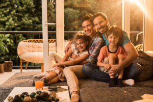 Imagem de uma família composta por quatro pessoas: um homem, uma mulher, uma menina e um menino. Eles estão abraçados no sofá em um ambiente com luz suave, provavelmente ao entardecer, e parecem felizes e acolhidos. O ambiente parece ser confortável e familiar, com uma janela ao fundo mostrando uma vista de árvores. A imagem transmite um sentimento de união e carinho familiar.