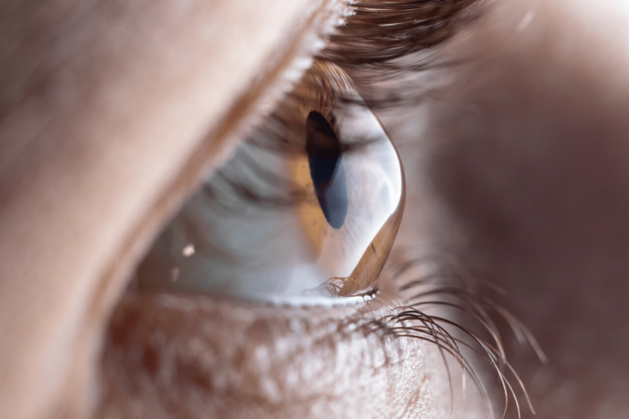A imagem mostra um close-up do olho de uma pessoa, destacando a córnea, a íris e a pupila. A luz refletida na córnea indica um alto nível de detalhe da estrutura ocular. A imagem foca principalmente na área ao redor da córnea, com a textura dos cílios e da pele ao redor do olho visíveis. Esse tipo de imagem é frequentemente utilizado para ilustrar a anatomia ocular em materiais educativos ou médicos.
