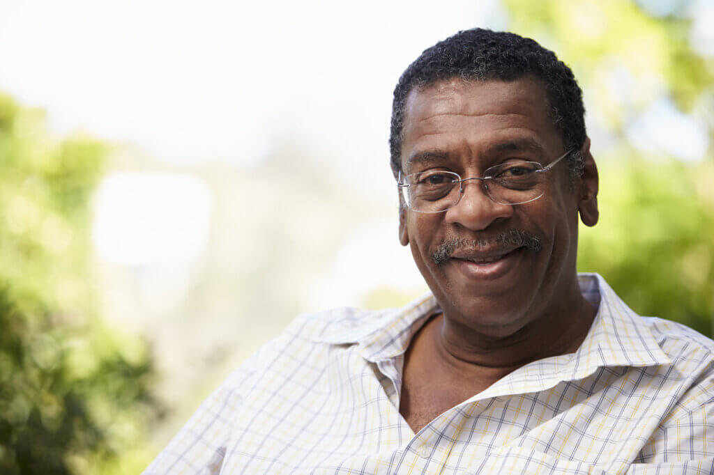 Imagem de um homem mais velho, com cabelo e bigode grisalhos, sorrindo e usando óculos. Ele está usando uma camisa de manga curta xadrez e parece estar em um ambiente ao ar livre, com uma luz suave e fundo desfocado. A expressão é amigável e tranquila, transmitindo uma sensação de felicidade e calma.