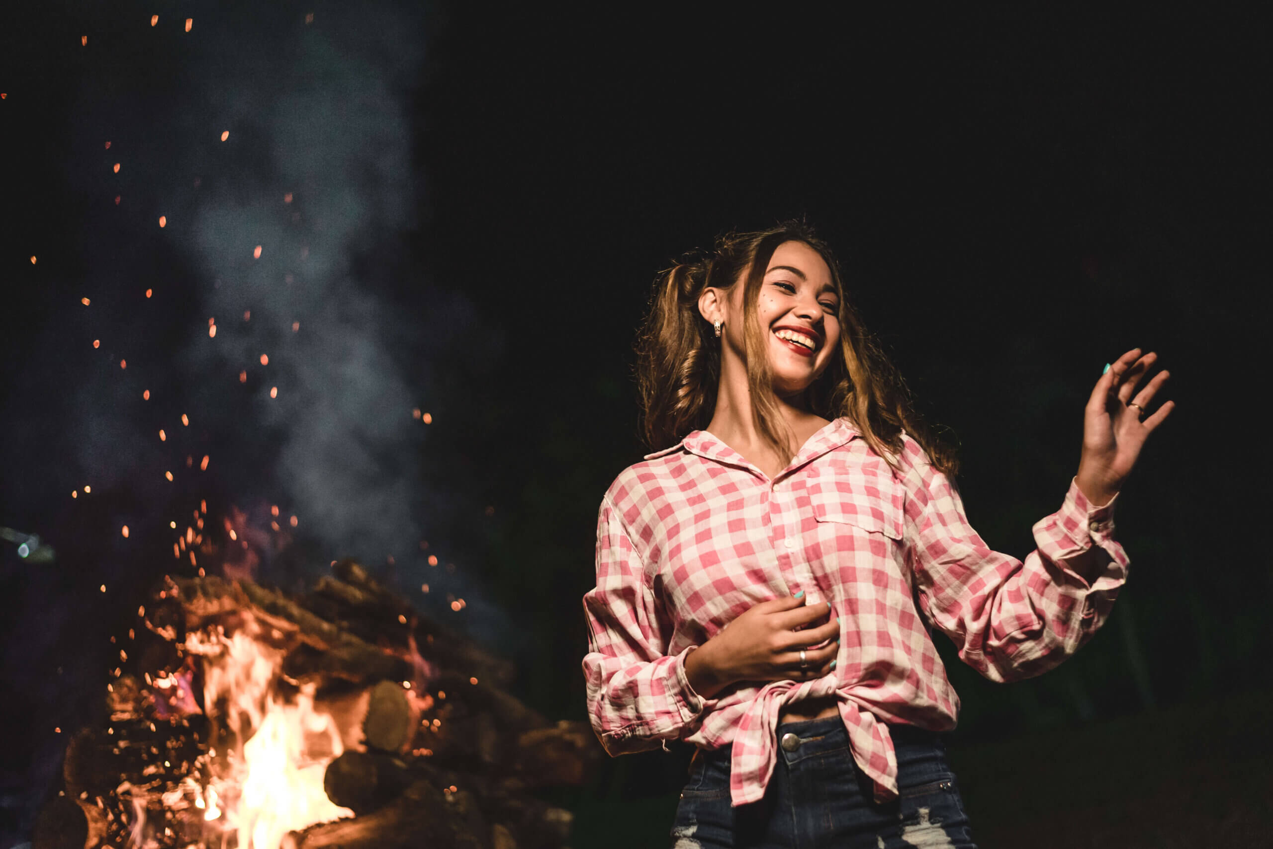 Imagem de uma jovem sorrindo enquanto dança ao lado de uma fogueira. Ela usa uma camisa xadrez rosa e branca, com o cabelo solto e uma expressão de alegria. O fundo é escuro, com faíscas iluminadas pela chama da fogueira, criando uma atmosfera acolhedora e festiva.