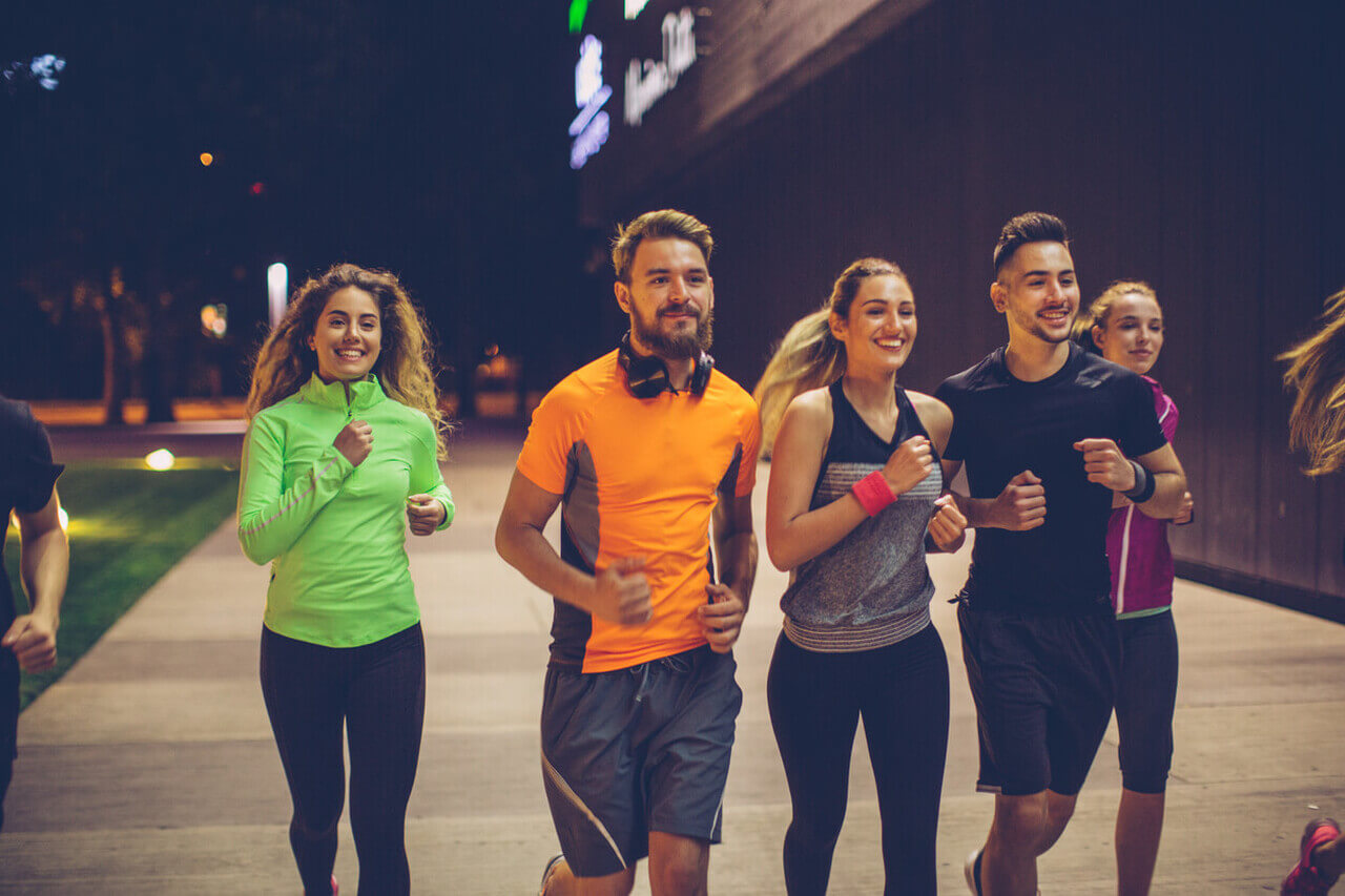 Imagem de um grupo de quatro pessoas correndo juntas à noite, com iluminação de rua ao fundo. Eles estão sorrindo e usando roupas esportivas coloridas e confortáveis. O ambiente sugere um momento de atividade física ao ar livre, provavelmente parte de um treino em grupo ou corrida casual.