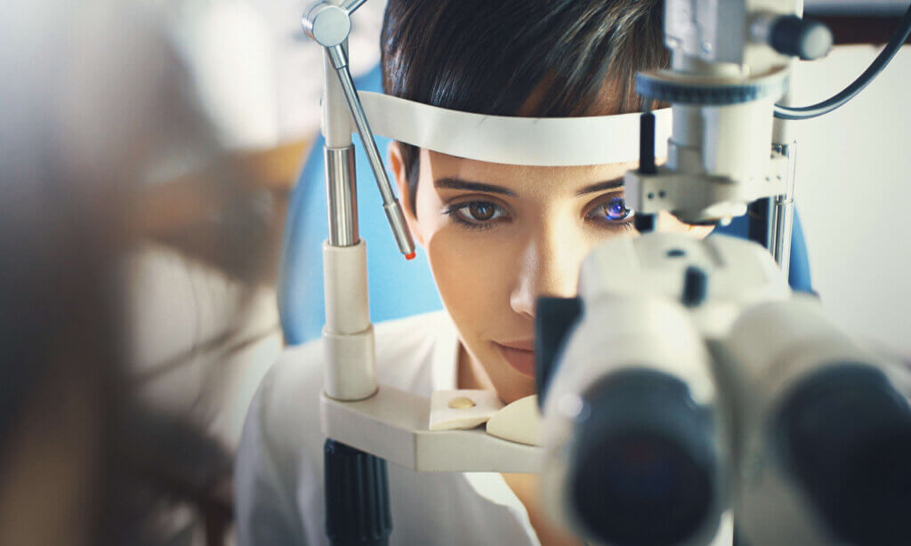 A imagem mostra uma mulher sendo examinada com um equipamento de oftalmologia, provavelmente um microscópio de fenda. Ela está focada no exame, com os olhos observando o aparelho, enquanto a luz do equipamento ilumina seu rosto. O fundo é desfocado, destacando a concentração e a técnica do exame oftalmológico.
