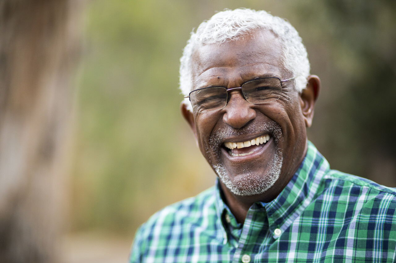 Homem idoso sorridente com cabelo grisalho e óculos, usando uma camisa xadrez verde, ao ar livre com fundo desfocado.