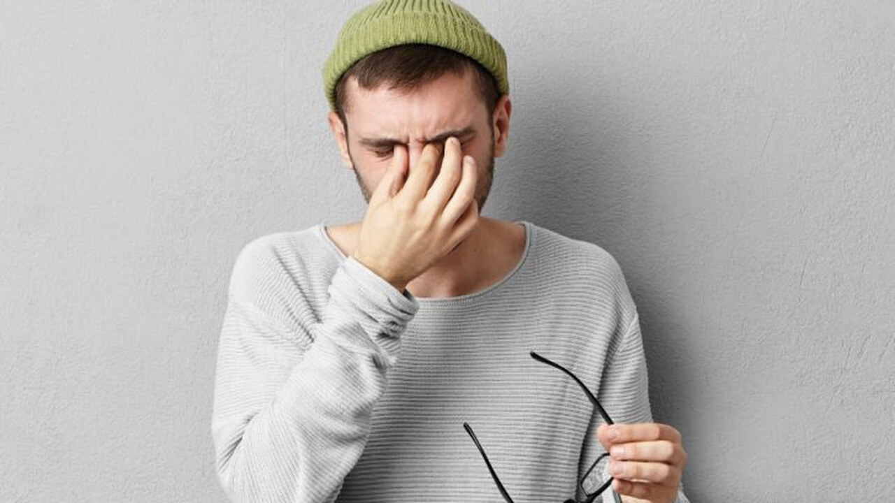
Um homem jovem com barba, usando uma touca verde e uma blusa cinza clara, esfrega os olhos com uma das mãos, enquanto segura óculos na outra mão, indicando cansaço visual ou irritação. Ele está em frente a uma parede cinza clara.