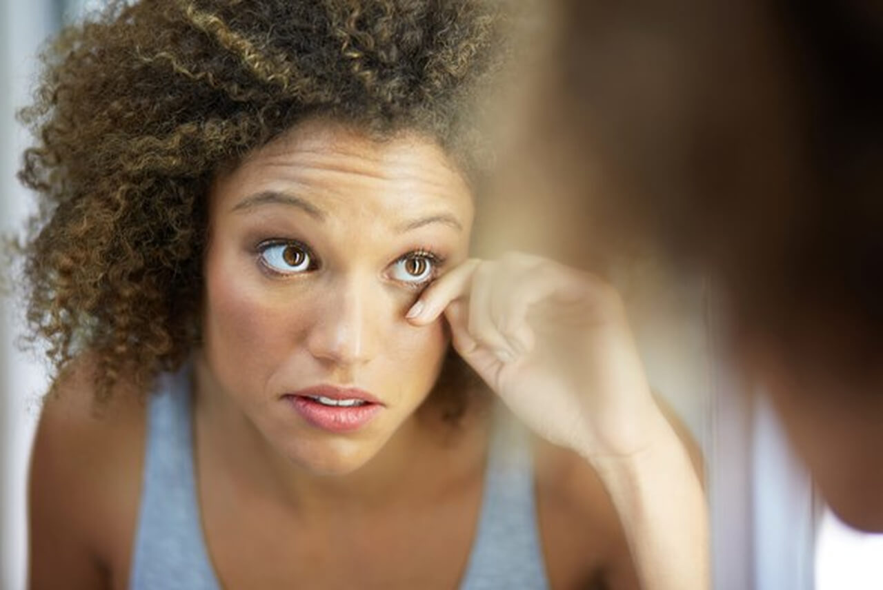 Uma mulher com cabelo cacheado e de pele escura olha para o seu reflexo em um espelho, puxando a pálpebra inferior de seu olho direito para examinar a parte branca do olho. Sua expressão facial indica preocupação.