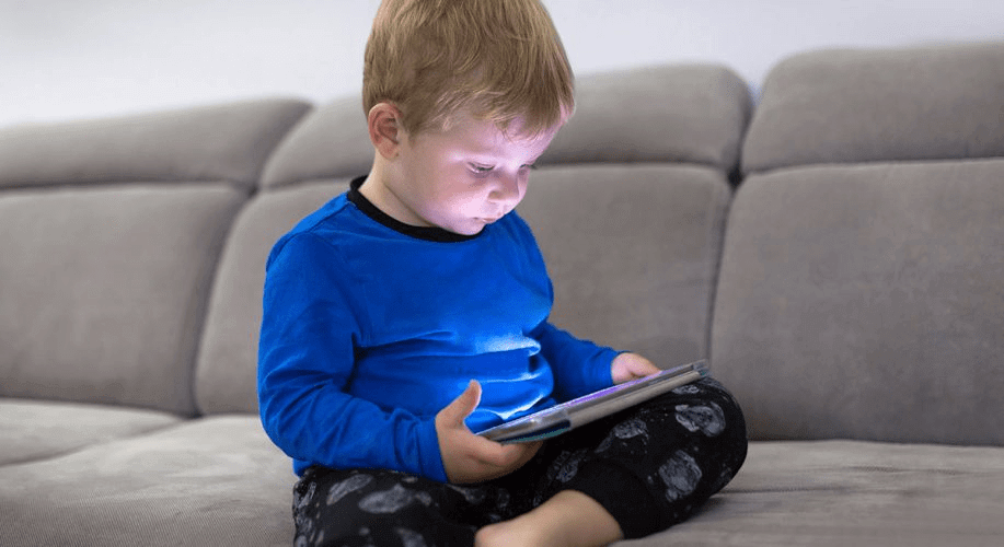 Imagem de um menino pequeno, sentado no sofá, olhando para uma tela de tablet. Ele está usando uma camiseta azul e calças de pijama com estampas de personagens. A luz da tela ilumina seu rosto, destacando sua atenção ao dispositivo, em um ambiente de iluminação suave.