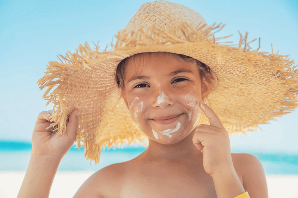 Imagem de uma criança sorrindo enquanto usa um chapéu de palha e aplica protetor solar no rosto, provavelmente na praia. O fundo é uma paisagem de céu azul e mar, transmitindo uma sensação de diversão e cuidado com a pele.