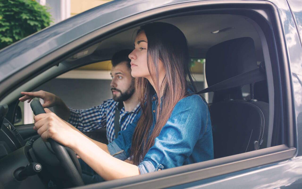 Imagem de um casal dentro de um carro, onde a mulher está dirigindo e o homem está ao seu lado, provavelmente oferecendo instruções ou acompanhando o trajeto. Ambos estão usando roupas casuais, e a mulher parece focada na direção. A cena ocorre durante o dia, com luz natural visível através das janelas do carro.