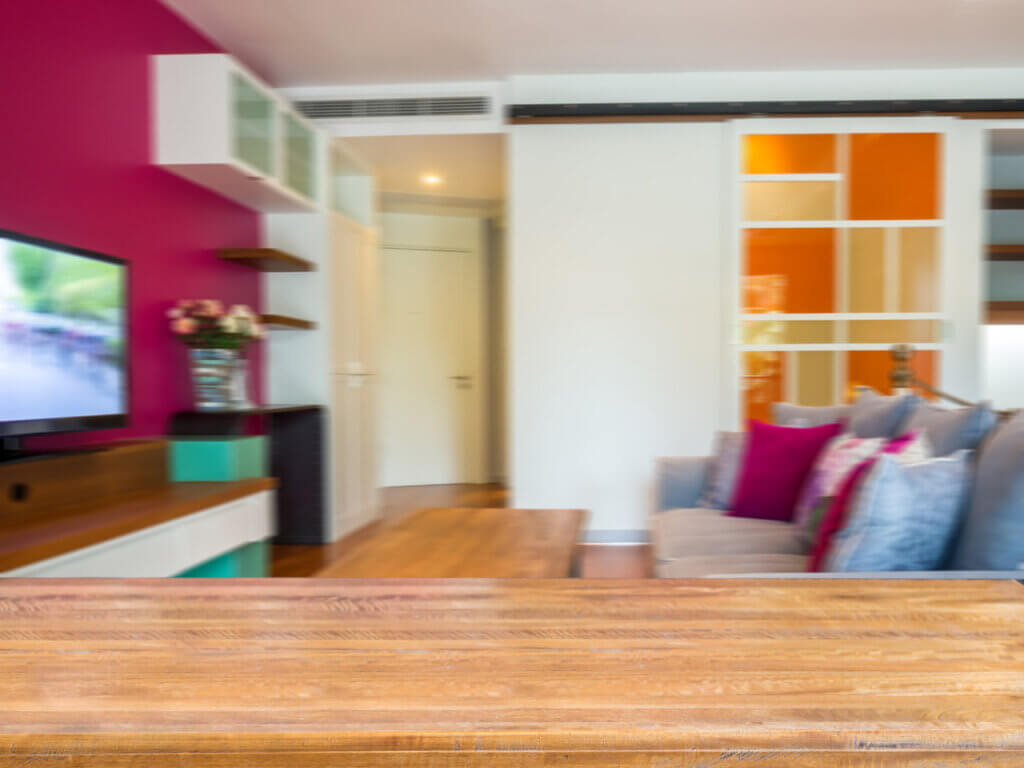 Imagem de uma sala de estar com móveis modernos e decoração colorida. O foco está em uma mesa de madeira em primeiro plano, enquanto ao fundo é possível ver um sofá com almofadas coloridas, uma parede com pintura em tons vibrantes de rosa e laranja, e uma TV em um rack. A imagem tem um leve desfoque, que dá a sensação de um ponto de vista capturado de um ângulo baixo.