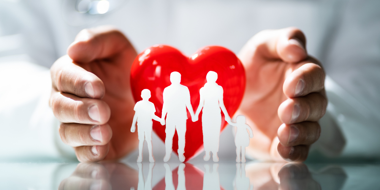 Mãos protegendo um coração vermelho com silhuetas brancas de uma família com quatro pessoas sobre uma superfície refletiva, representando cuidado e proteção familiar.