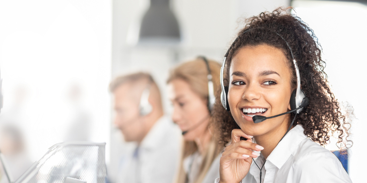 
Uma jovem sorridente com cabelo crespo e fone de ouvido com microfone, possivelmente trabalhando em um call center, com outras pessoas desfocadas ao fundo. Ela está vestindo uma camisa branca.