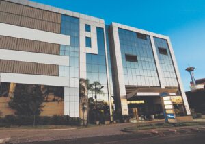  Um edifício moderno de escritórios, com fachada de vidro e detalhes em branco e cinza, sob um céu azul. Há folhagens na parte inferior e uma torre de telecomunicações distante no lado direito.